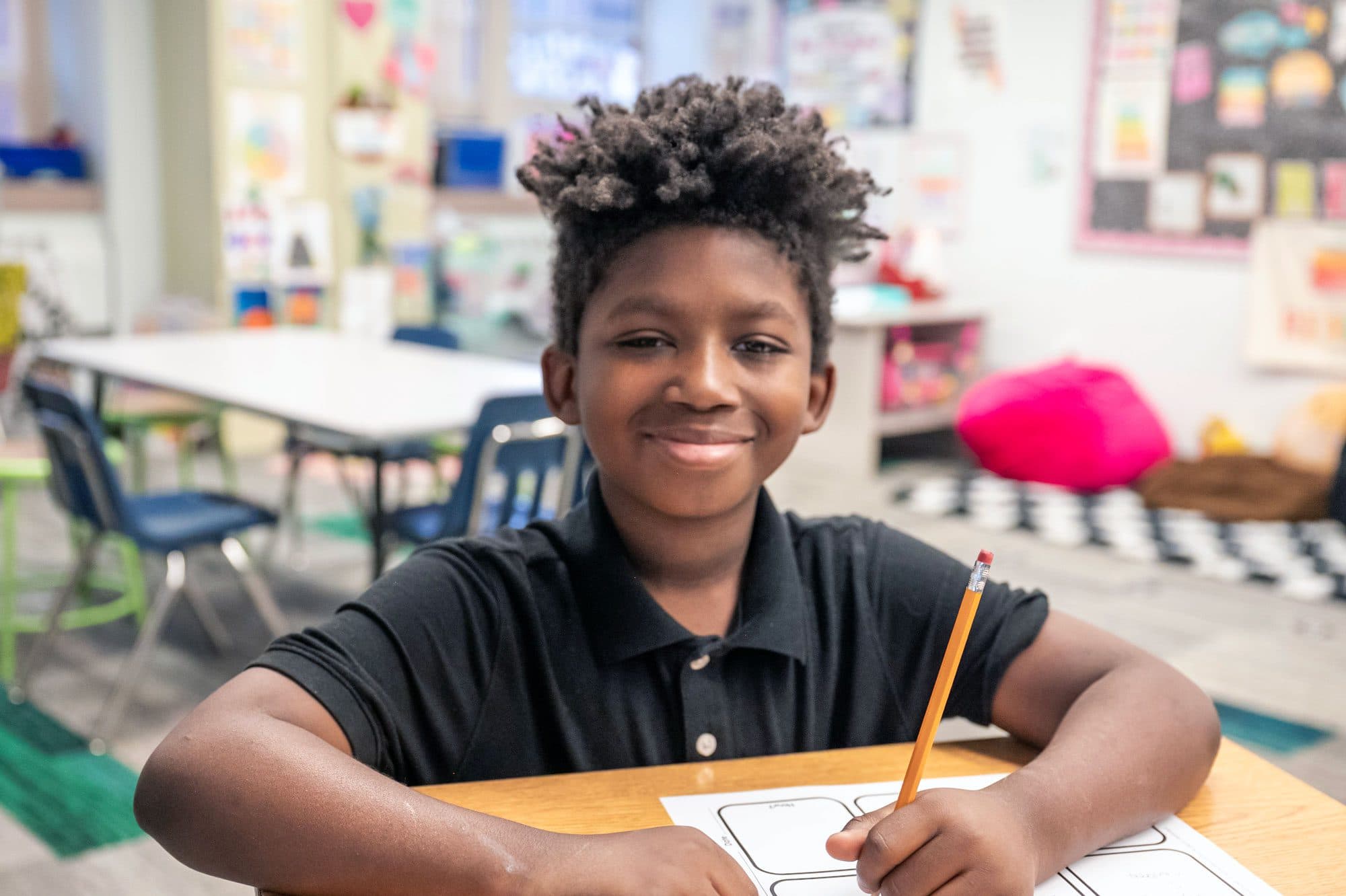 Smiling student writing on a worksheet at a classroom desk.