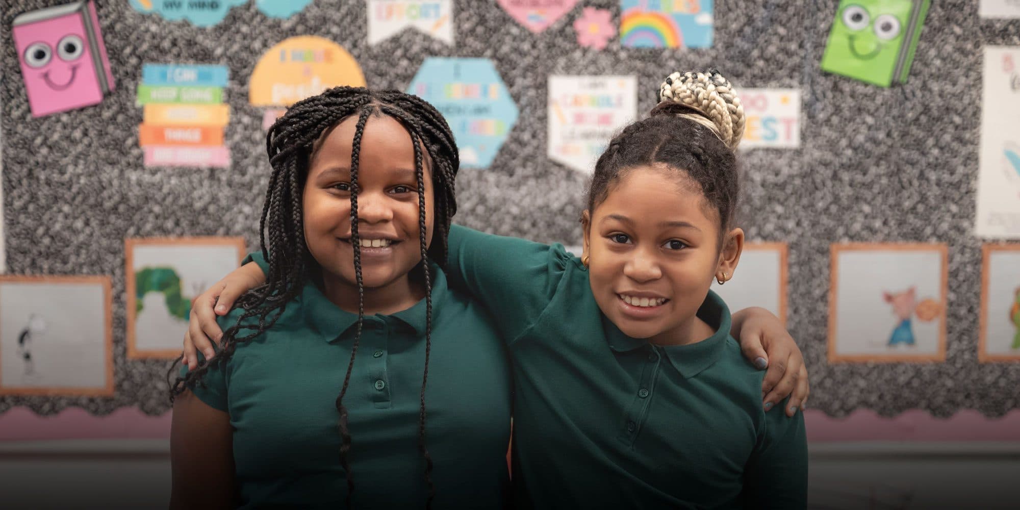 Two smiling students in green uniform shirts standing arm in arm in front of a classroom bulletin board.