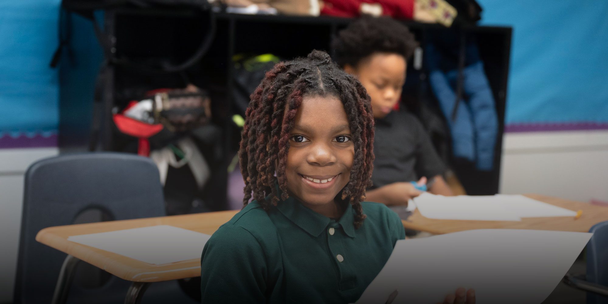 Smiling student in a green polo holding paper at a classroom desk.