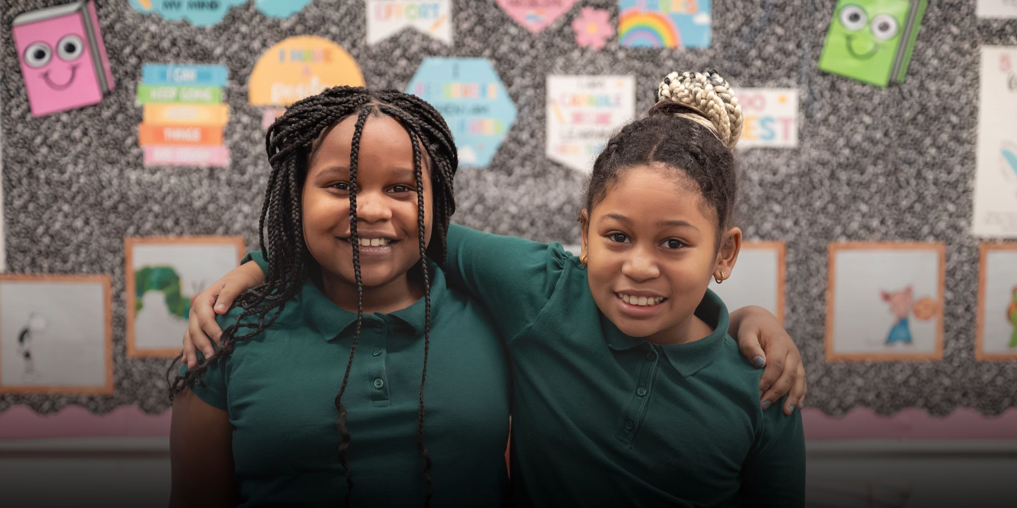 Two smiling students in green uniform shirts standing arm in arm in front of a classroom bulletin board.